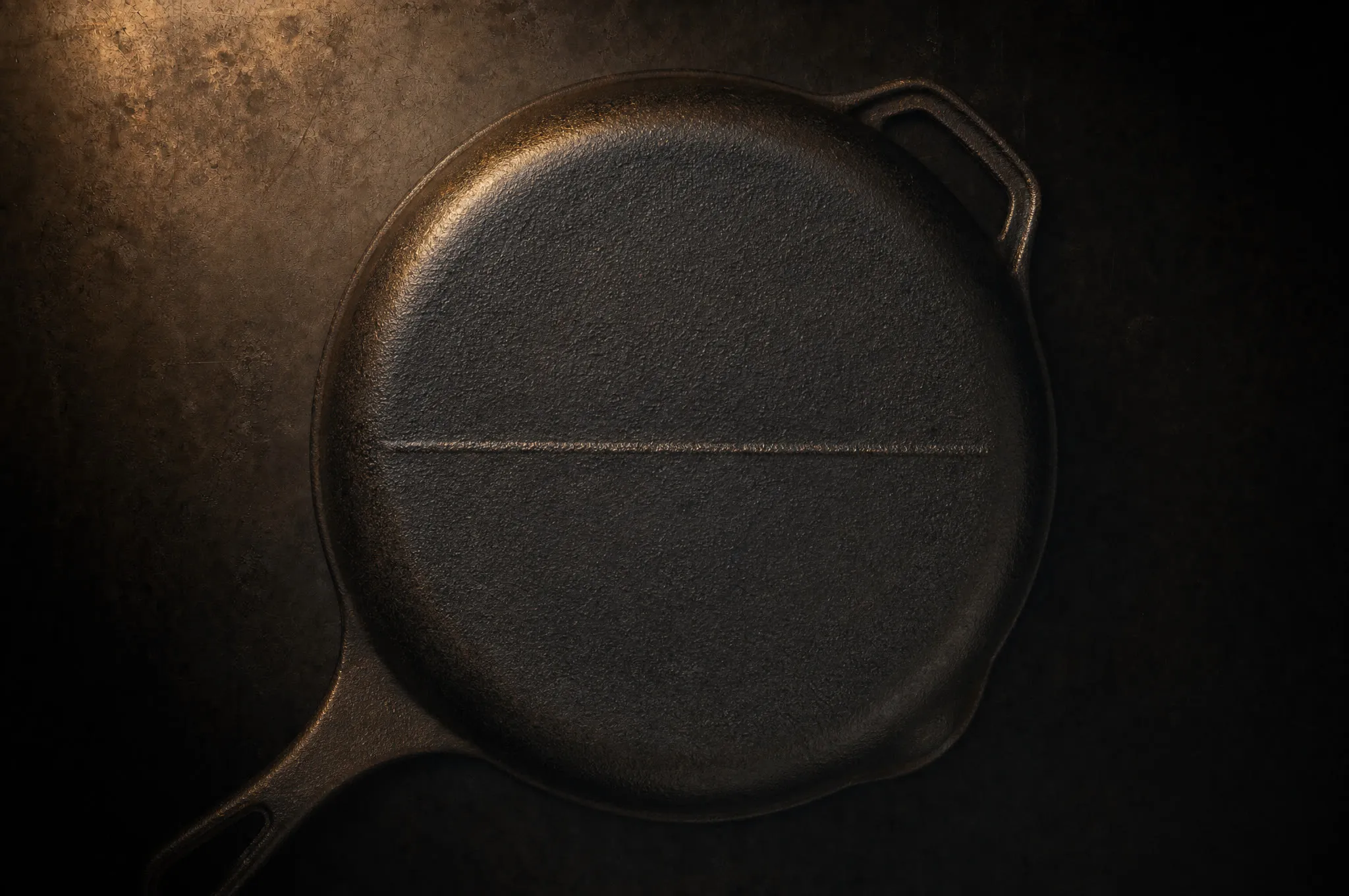 Underside of an antique cast iron skillet in raking light showing a thin raised gate mark scar across the bottom